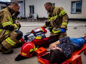 na noszach lezy poszkodowana kobieta, obok strażacy udzielają jej pierwszej pomocy medycznej