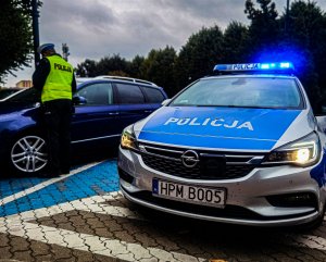 radiowóz, obok stoi policjant i sprawdza czy pojazd pozostawiony na miejscu parkingowym dla osób niepełnosprawnych pozostawioną ma na podszybiu kartę inwalidzką