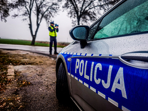 bok radiowozu, w tle policjant na drodze mierzy prędkość